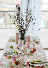 Decorative festive table setting with floral tablecloth, egg, dish and golden cutlery, glass for wine, willow branch. Happy easter holidsy concept.