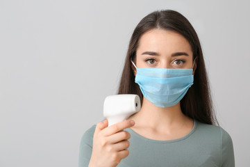 Young woman with medical mask and infrared thermometer against grey background