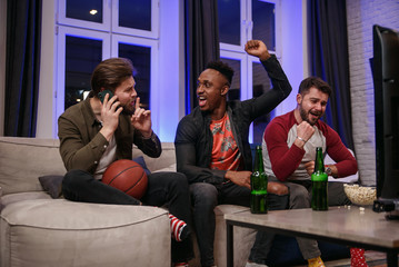 Fierce noisy likable 30s multiracial male friends encouraging with shouts their favourite football team and interfering their male friend to have mobile conversation