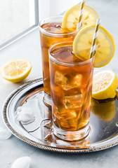 Two frosty glasses of ice tea with lemon slices on a metal tray against a window sill.