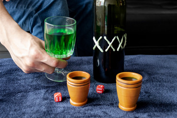 Close up to a man holding a cup of green homemade liquor with two wooden cups and green artesanal alcohol bottle and double six red dices. Party and liquor games concept
