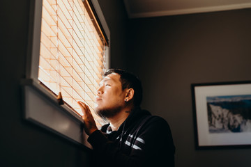 Asian man in isolation looking through window blinds. Man forced to stay inside the house as a...