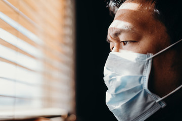 Asian man in isolation looking through window blinds. Man in medical mask forced to stay inside the...
