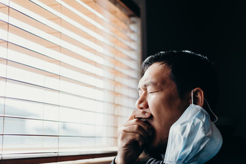 Asian man in isolation looking through window blinds and cough. Man in medical mask forced to stay...