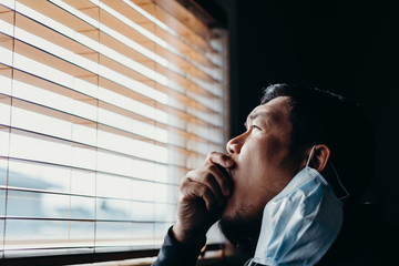 Asian man in isolation looking through window blinds and cough. Man in medical mask forced to stay...