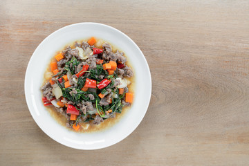 top view of stir fried minced beef with holy basil in a ceramic dish on wooden table. hot and spicy thai style food menu.