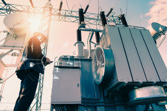 Businessman wearing a black suit, standing looking at a large power transformer with orange sunlight and white sky to be background, Concept about business people who want to invest in energy