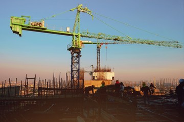 The construction of a rooftop high rise building, workers are tied with steel bars for concrete pouring, surveying the site and cranes are lifting or moving metal at the construction site. Sunset