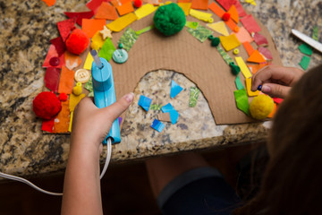 Child putting object on a rainbow collage while holding a hot glue gun