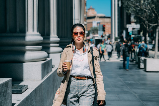 Woman outdoors listening to music with headphones and drinking to go paper cup of coffee. confident asian chinese college girl on way to university in city street. elegant lady in sunglasses walking