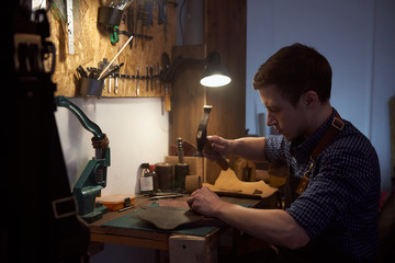 The craftsman works with the hammer and the awl to punch small holes on genuine leather for the seam. Young leatherman works with natural leather using craft tools. A small business of a craftsman.