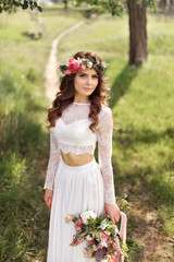 Obraz na płótnie Canvas The bride in rustic style with a beautiful wreath on the head of flowers with a bouquet in hands standing in a park in white dress