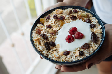 Healthy breakfast, a woman holds in her hands a bowl of oatmeal and yogurt with raspberry, nuts and dried raisins