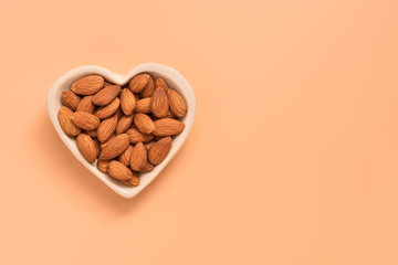 Almond nut in a heart shape bowl on a pastel pink background. Healthy eating concept. Top view,...