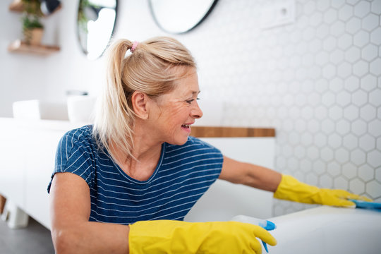 Senior woman with gloves cleaning bathroom indoors at home.