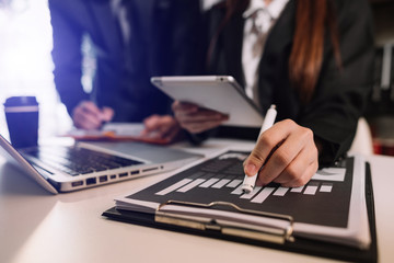 Business documents on office table with smart phone and laptop and two colleagues discussing data in the background in morning light
