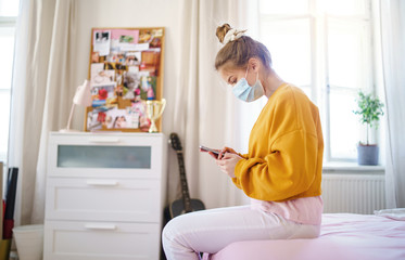 Young female student with face mask sitting on bed, using smartphone.
