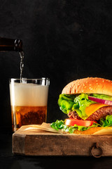 Burger and beer. Hamburger with beef, cheese, onion, tomato, and green salad, a side view on a dark background with copy space. Selective focus
