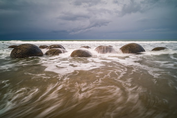 Waves washing against the mysterious Moeraki Boulders with dark storm clouds overhead at Koekohe beach on the Otago coast of New Zealand. These amazing round stones were formed millions of years ago