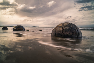 Waves washing against the mysterious Moeraki Boulders with dark storm clouds overhead at Koekohe beach on the Otago coast of New Zealand. These amazing round stones were formed millions of years ago