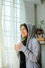 Young adult happy muslim woman enjoying hot chocolate drinks by the window while wearing hoodie
