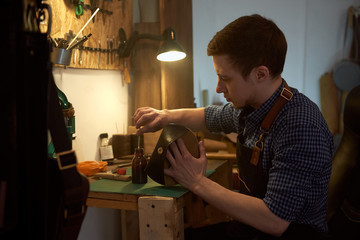Young master paints the edges of the leather pattern for future product. The craftsman working with brown leather using craft tools. Handmade leather goods manufacturing. Leathermaker's work desk.