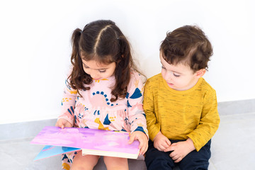 Two cute siblings reading book in home. Toddlers girl and boy having fun together. Happiness is in small things, slow living, education in home concept.