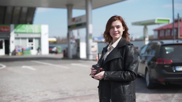 service, satisfied female client drinks coffee at a gas station while her car is fueled with gasoline, fuel prices