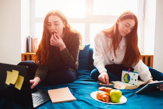 Young women doing homework in a dormitory bed near the window. There are notebooks, food books, a tablet and laptop and documents