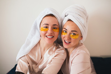 Two young women having fun with patches under their eyes. Two friends in towels and pajamas have a fun spa party together in home.
