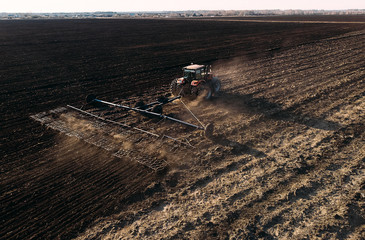 Aerial view of tractor plows the field in sunset, sunrise, raising dust, and behind it fly birds. Farm tractor with harrow plow preparing land for sowing. Agriculture industry, cultivation of land.