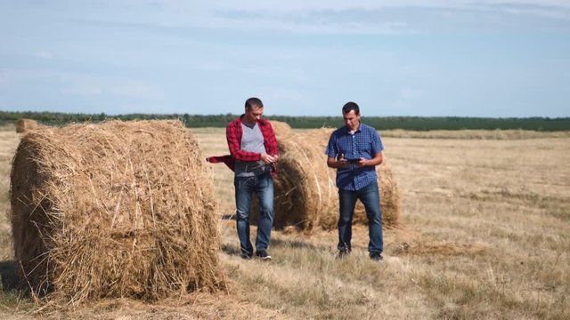 Teamwork. Two farmers study straw bales. Male agronomists in jeans and shirts in a wheat field after harvesting, studying bales, rolled straw with a tablet in their hands. Teamwork success