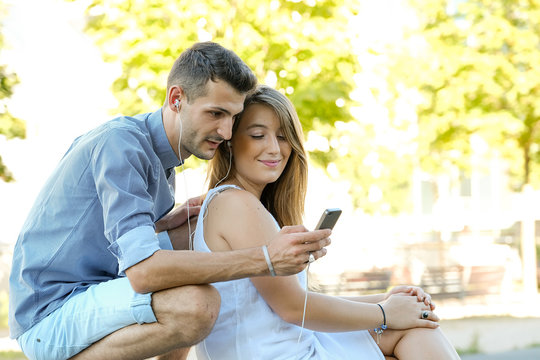 Happy young students couple using digital tablet and cellphone with earpeaces outside on summer season with green city background
