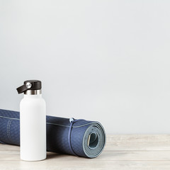 Rolled blue yoga mat and white metal water bottle flask on grey wooden surface. Gender neutral fitness and exercise concept and hydration with copy space