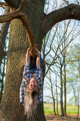 Junge Frau hängt lachend Kopfüber an einem Ast von einem dicken Baum. Ansicht von vorne. Standort: Deutschland, Nordrhein Westfalen, Hoxfeld