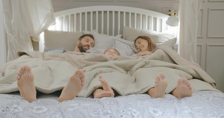 Young happy mother, father and little son lying in bed and talking in morning. Cheerful parents and child chatting relaxing in bed under blanket with bare feet on foreground