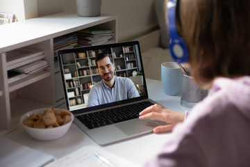 Student girl wearing headphones sitting at desk watching educational webinar, listen teacher during distant lesson via videoconference, view over woman shoulder laptop screen male tutor teach learner