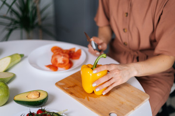 Close-up of hand of woman cutting fresh bell pepper using kitchen knife on wooden cutting board. Young woman cutting yellow bell pepper with a knife for vegetable salad. Concept of healthy eating.