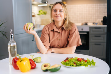 Attractive woman sits at table in modern kitchen, holds in her hand fresh fruit, on the table avocado, bell pepper, green salad, olive oil. Concept of healthy eating.