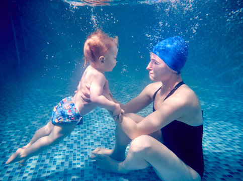 Side view of young woman in swimsuit holding adorable child while sitting on swimming pool floor. Loving mother teaching baby to swim underwater. Concept of family, motherhood and active lifestyle.