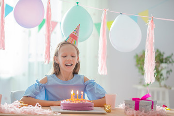 Caucasian girl is dreamily smiling and looking at birthday rainbow cake. Festive colorful...
