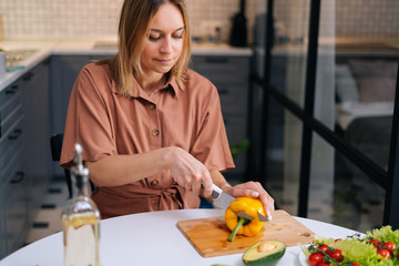 Young woman cutting fresh yellow bell pepper with a knife for vegetable salad in modern kitchen. Smiling slim vegetarian female prepare healthy vegan dinner meal. Concept of healthy eating.