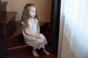 Fototapeta premium Sad little girl sits on the steps of the stairs inside the house and looks out the window with longing