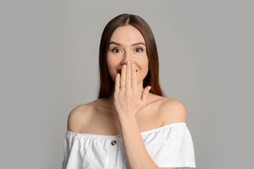 Emotional young woman wearing beautiful engagement ring on grey background