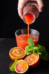 Top view of hand serving blood orange juice in glass cup, with half orange and mint, on black...
