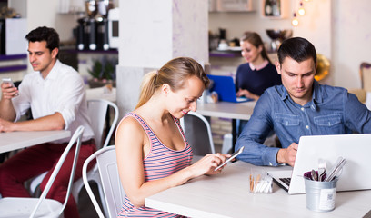 Young smiling woman with boyfriend busying with device in modern cafe