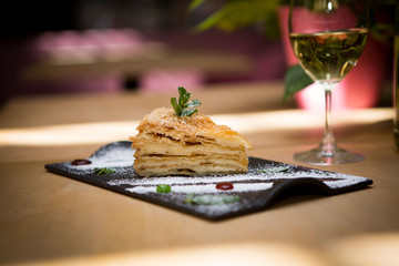 On the table is a glass of white wine and a portion of delicious cake on a black plate sprinkled with powdered sugar, next to which are appliances for food.