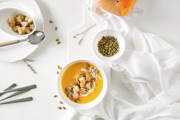Pumpkin soup, a piece of pumpkin, seeds and crackers lie on a white table. Nearby is a white towel and serving utensils. 
