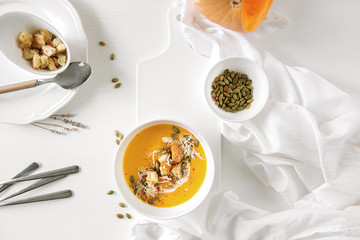 Pumpkin soup, a piece of pumpkin, seeds and crackers lie on a white table. Nearby is a white towel and serving utensils. 

