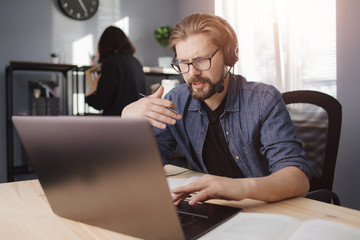 Competent businessman in domestic outfit having video conference while sitting at home. Bearded man in eyeglasses using laptop and headset for remote work.